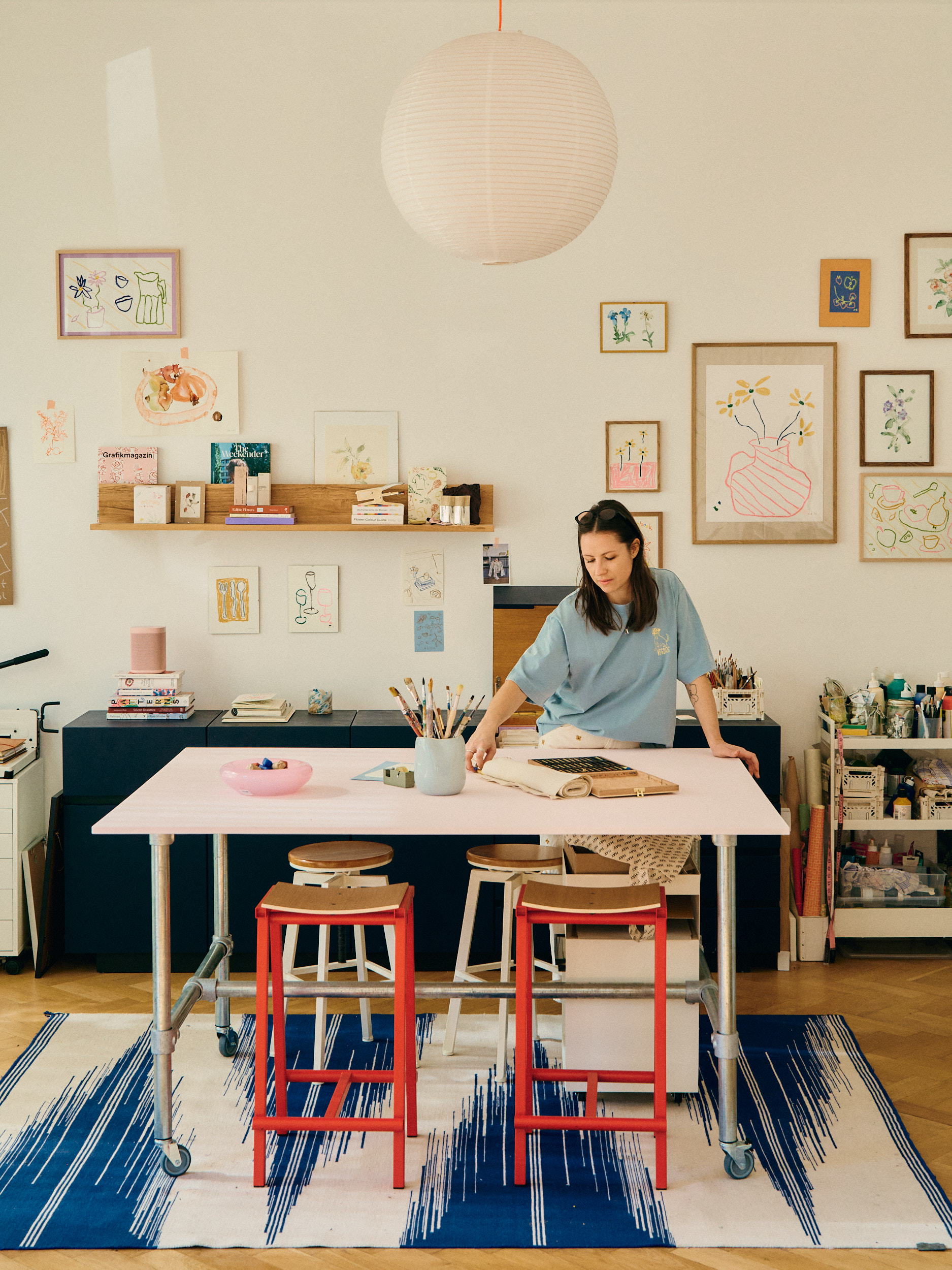 Heller, liebevoll eingerichteter Kreativarbeitsraum mit großem Arbeitstisch in der Mitte. Eine Frau steht am Tisch und arbeitet mit Skizzenbuch und Materialien. Auf dem Tisch liegen Pinsel, Farben und Bücher, darunter stehen Hocker. Die Wände sind mit vielen kleinen, farbenfrohen Illustrationen und gerahmten Zeichnungen dekoriert. Regale und Aufbewahrungsboxen mit weiteren Materialien unterstreichen die kreative, persönliche Atelier-Atmosphäre.