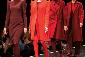 Vier Modells laufen auf einem Catwalk in verschiedenen Rottönen – von Bordeaux bis leuchtendem Rot. Alle tragen markante, breite Sonnenbrillen und strukturierte Mäntel oder Kleider mit betonten Schultern.