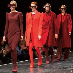 Vier Modells laufen auf einem Catwalk in verschiedenen Rottönen – von Bordeaux bis leuchtendem Rot. Alle tragen markante, breite Sonnenbrillen und strukturierte Mäntel oder Kleider mit betonten Schultern.