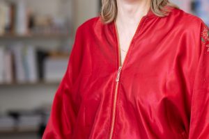 Porträt einer lächelnden Frau mit blondem, schulterlangem Haar in einem hellen Büro oder Atelier. Sie trägt eine glänzende rote Jacke mit floraler Stickerei am Ärmel und steht entspannt mit den Händen in den Taschen vor einem unscharfen Hintergrund mit Bücherregal und großformatigem Bild oder Bildschirm in Lila- und Rosatönen.