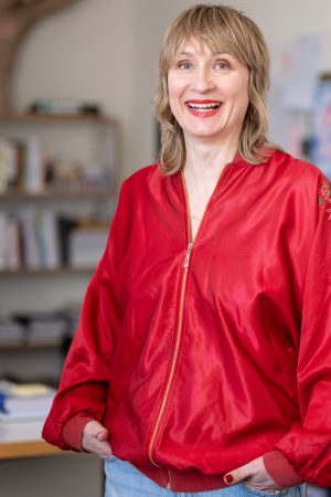 Porträt einer lächelnden Frau mit blondem, schulterlangem Haar in einem hellen Büro oder Atelier. Sie trägt eine glänzende rote Jacke mit floraler Stickerei am Ärmel und steht entspannt mit den Händen in den Taschen vor einem unscharfen Hintergrund mit Bücherregal und großformatigem Bild oder Bildschirm in Lila- und Rosatönen.