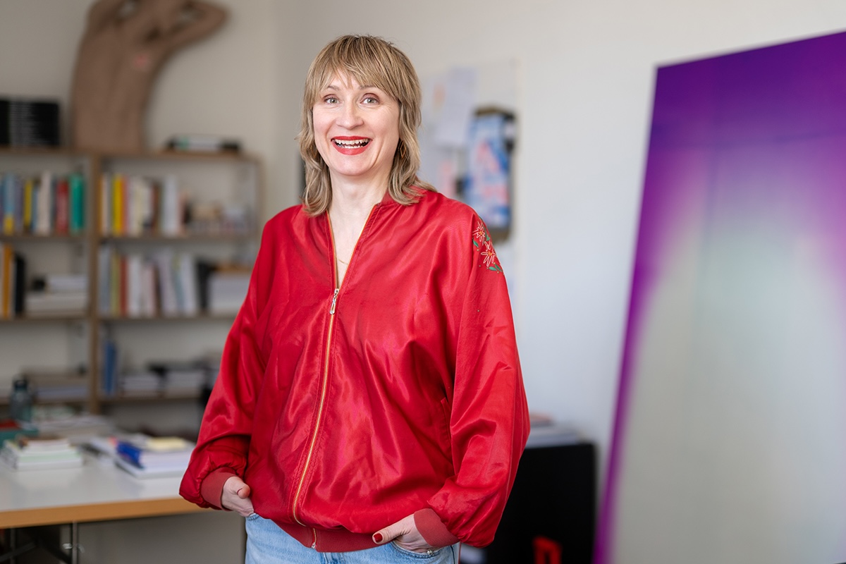 Porträt einer lächelnden Frau mit blondem, schulterlangem Haar in einem hellen Büro oder Atelier. Sie trägt eine glänzende rote Jacke mit floraler Stickerei am Ärmel und steht entspannt mit den Händen in den Taschen vor einem unscharfen Hintergrund mit Bücherregal und großformatigem Bild oder Bildschirm in Lila- und Rosatönen.