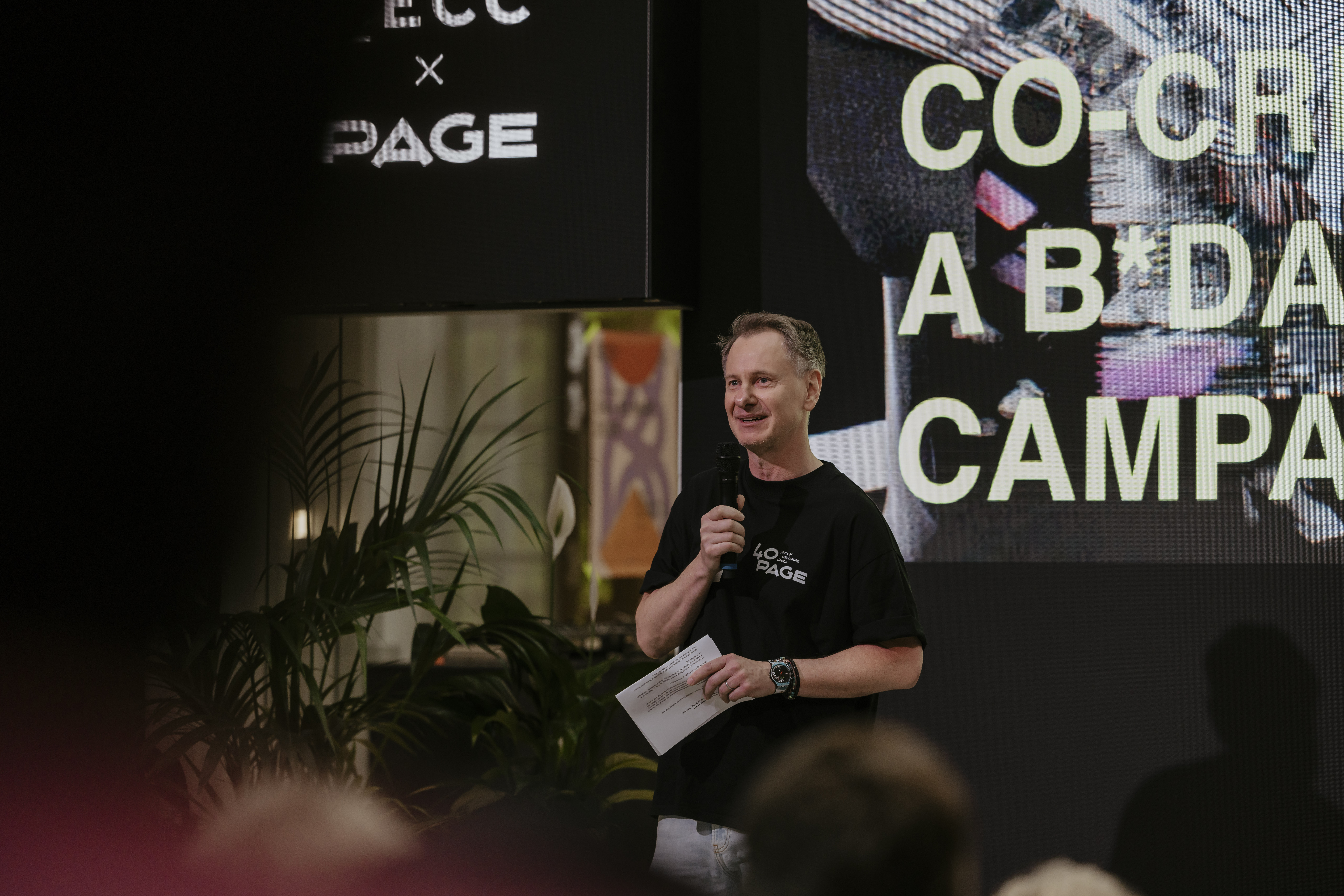 Mann steht auf einer Bühne und spricht in ein Mikrofon, während er in der anderen Hand Notizen hält. Hinter ihm ist eine große Leinwand mit der Aufschrift „BECC x PAGE“ sowie dem Titel „Co-Creation Birthday Campaign“ zu sehen. Er trägt ein schwarzes T-Shirt mit „40 PAGE“-Logo. Im Vordergrund sind unscharf sitzende Zuhörer zu erkennen, links steht eine große Zimmerpflanze, die dem Event-Setting eine lockere Atmosphäre verleiht.