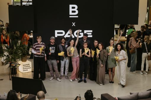 Gruppe von etwa zehn Personen steht auf einer Bühne vor einer großen schwarzen LED-Wand mit der Aufschrift „B x PAGE“. Sie halten gelbe Blumen in der Hand, einige lächeln in die Kamera, eine Person in der Mitte hebt jubelnd beide Arme. Das Setting wirkt wie eine Preisverleihung oder ein Community-Event. Im Hintergrund stehen weitere Gäste im Gespräch, links ist ein Podest mit der Aufschrift „munich creative business week“ sowie Pflanzen und Getränke zu sehen. Im Vordergrund sitzen Zuschauer und applaudieren.