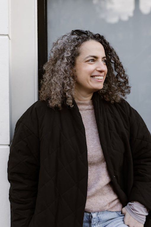 Porträt einer lächelnden Person mit schulterlangen dunklen Locken, die an eine Hauswand gelehnt steht. Sie trägt eine schwarze gesteppte Jacke über einem hellen Strickpullover und eine helle Jeans. Die Person blickt nach rechts aus dem Bild, während im Hintergrund ein großes Fenster und eine helle Außenwand zu sehen sind. Die Aufnahme wirkt natürlich, freundlich und entspannt.