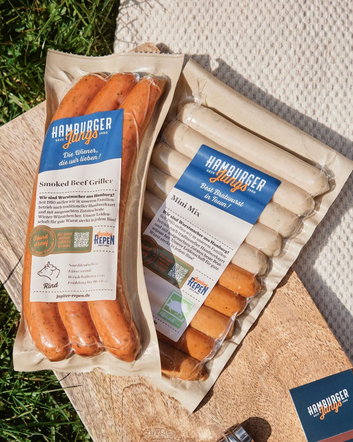 Zwei vakuumverpackte Würstchen-Packungen der Marke „Hamburger Jungs“ liegen auf einem Holzbrett im Freien. Eine Packung enthält „Smoked Beef Griller“, die andere eine „Mini Mix“-Auswahl mit hellen und dunkleren Würstchen. Die Verpackungen sind mit blauen Etiketten gestaltet und von Gras und Sonnenlicht umgeben.