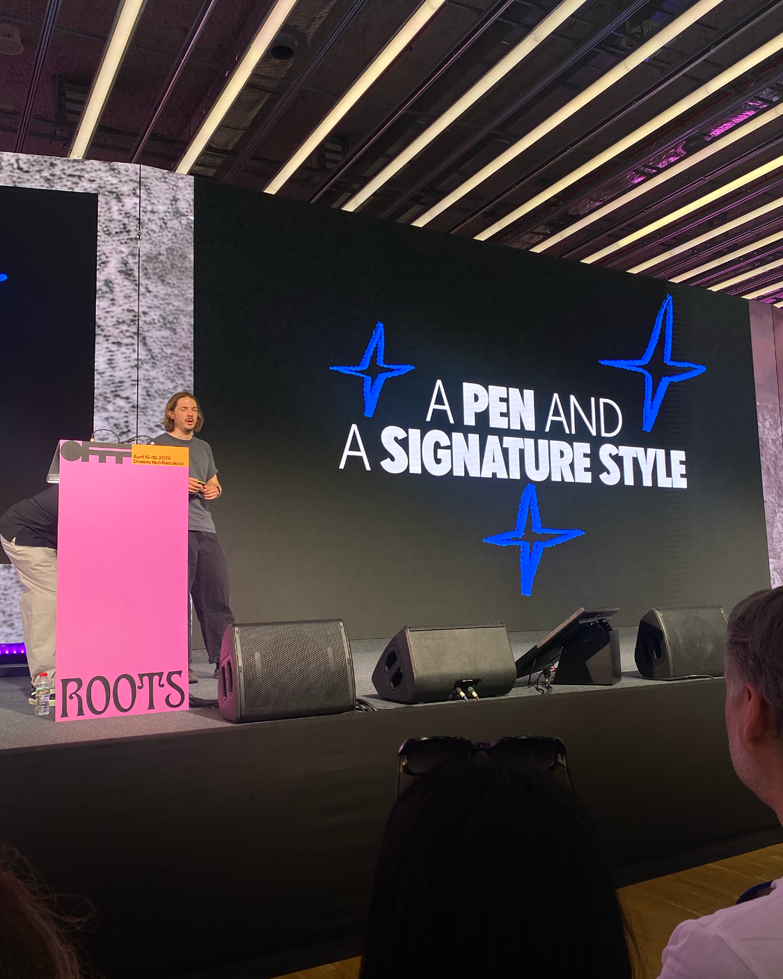 Ein Mann steht auf einer Bühne neben einem pinken Rednerpult mit der Aufschrift „ROOTS“ und hält einen Vortrag. Hinter ihm zeigt eine große Leinwand den Text „A PEN AND A SIGNATURE STYLE“ mit grafischen Elementen. Im Vordergrund sind Teile des Publikums zu sehen, die zur Bühne blicken.