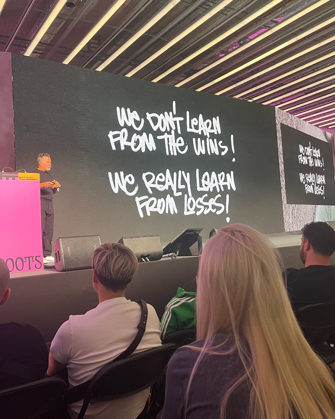 Ein Mann steht auf einer Bühne neben einem pinken Rednerpult und spricht vor Publikum. Hinter ihm zeigt eine große Leinwand den handschriftlich wirkenden Text: „We don’t learn from the wins! We really learn from losses!“. Im Vordergrund sitzen Zuhörer und verfolgen den Vortrag.