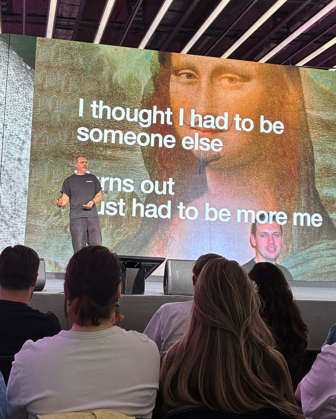 Ein Mann steht auf einer Bühne und hält einen Vortrag vor Publikum. Hinter ihm ist eine große Leinwand mit einem Ausschnitt der Mona Lisa und dem Text „I thought I had to be someone else – turns out I just had to be more me“. Im Vordergrund sitzen Zuhörer und blicken zur Bühne.