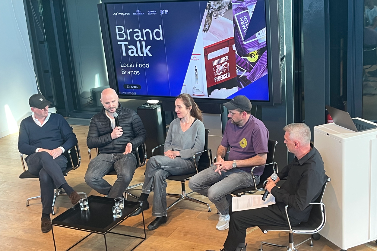 Fünf Personen sitzen auf einer Bühne in einer Diskussionsrunde („Brand Talk – Local Food Brands“) und sprechen miteinander. Eine Person hält ein Mikrofon, während die anderen zuhören. Hinter ihnen zeigt ein großer Bildschirm den Titel der Veranstaltung und visuelle Beispiele von Marken. Im Vordergrund stehen ein kleiner Tisch mit Wassergläsern sowie ein Moderator mit Mikrofon.