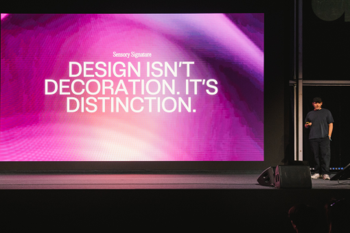 Ein Mann steht auf einer Bühne und hält einen Vortrag neben einer großen, leuchtenden Leinwand. Darauf ist ein abstraktes, pinkfarbenes Motiv zu sehen sowie der Text: „Design isn’t decoration. It’s distinction.“ Im abgedunkelten Raum richtet sich die Aufmerksamkeit des Publikums auf die Präsentation.