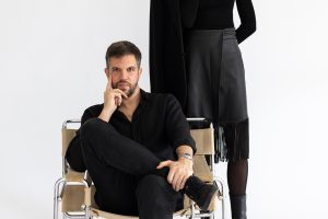 Zwei kreativ wirkende Personen in einem minimalistischen Studio-Setting. Ein Mann sitzt entspannt auf einem modernen Freischwinger-Stuhl und blickt direkt in die Kamera, während eine Frau hinter ihm steht und ein schwarzes Sakko über der Schulter trägt. Beide sind vollständig in Schwarz gekleidet, wodurch ein eleganter, reduzierter Look entsteht. Der helle Hintergrund und das klare Styling vermitteln Professionalität, Ruhe und gestalterisches Selbstbewusstsein.