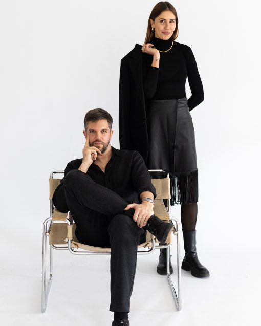 Zwei kreativ wirkende Personen in einem minimalistischen Studio-Setting. Ein Mann sitzt entspannt auf einem modernen Freischwinger-Stuhl und blickt direkt in die Kamera, während eine Frau hinter ihm steht und ein schwarzes Sakko über der Schulter trägt. Beide sind vollständig in Schwarz gekleidet, wodurch ein eleganter, reduzierter Look entsteht. Der helle Hintergrund und das klare Styling vermitteln Professionalität, Ruhe und gestalterisches Selbstbewusstsein.
