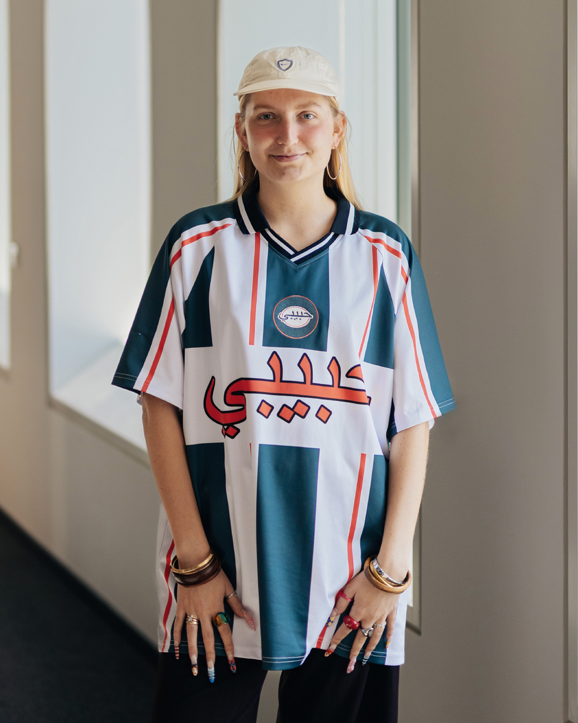 Eine junge Person mit heller Kappe und großen Creolen steht in einem hellen Flur vor Fenstern und blickt direkt in die Kamera. Sie trägt ein locker geschnittenes, sportliches Trikot mit grafischem Muster und auffälliger Schrift, dazu mehrere Ringe und Armbänder, die den lässigen Stil unterstreichen.