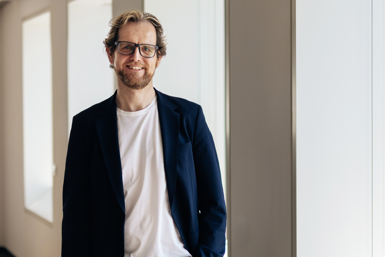Ein Mann mit Brille und leichtem Bart steht in einem hellen Flur vor großen Fensteröffnungen und lächelt in die Kamera. Er trägt ein dunkles Sakko über einem weißen T-Shirt und wirkt entspannt und zugänglich.