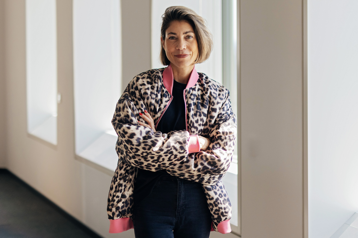 Eine Frau mit kurzem, seitlich gestyltem Haar steht in einem hellen Flur vor großen Fensteröffnungen, die Arme selbstbewusst verschränkt. Sie trägt eine gemusterte Jacke im Leo-Look mit auffälligem pinken Kragen über einem dunklen Outfit und blickt ruhig und direkt in die Kamera.