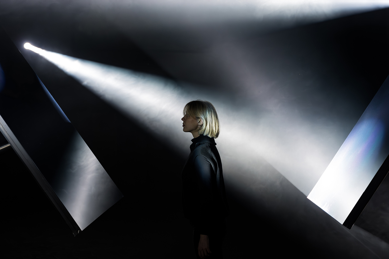 Eine Person im Profil steht in einem dunklen Raum und wird von starken, gerichteten Lichtstrahlen beleuchtet, die durch Nebel sichtbar werden. Die Szene wirkt inszeniert und künstlerisch, mit geometrischen Flächen und Lichtkegeln, die eine dramatische, fast futuristische Atmosphäre erzeugen.