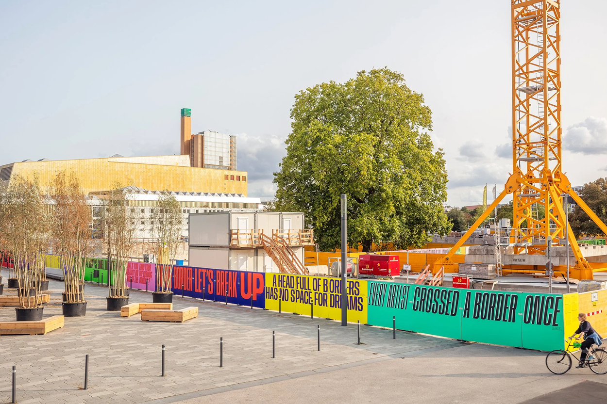 Städtische Baustelle mit Kran und temporären Gebäuden, umgeben von einem langen, farbenfrohen Bauzaun mit großen typografischen Botschaften. Im Vordergrund stehen junge Bäume in Pflanzkübeln, rechts fährt eine Person mit dem Fahrrad vorbei, während im Hintergrund ein großes Gebäude und ein Baum zu sehen sind.