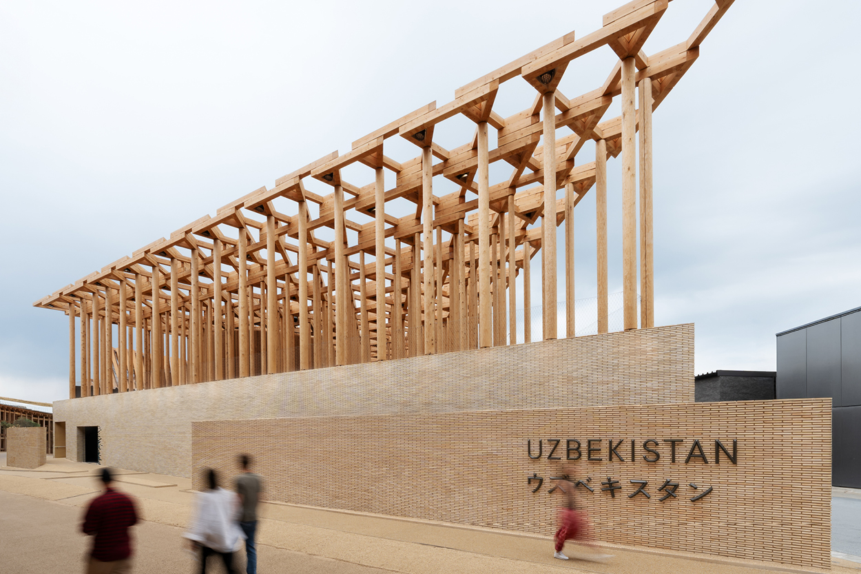 Moderner Pavillon mit offener Holzkonstruktion und filigranen Säulen steht vor einer langen Ziegelwand mit der Aufschrift „UZBEKISTAN“. Im Vordergrund bewegen sich einige unscharfe Besucher, während der Himmel grau und bedeckt ist.