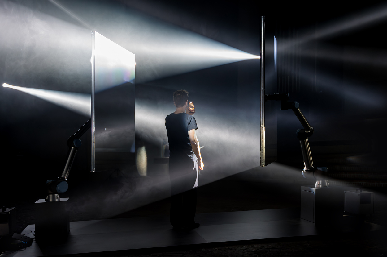 Eine Person steht in einem dunklen Raum zwischen technischen Installationen und wird von mehreren starken Lichtstrahlen angestrahlt. Sie hält ein Smartphone in der Hand, während um sie herum Nebel und Lichtkegel eine futuristische, experimentelle Atmosphäre erzeugen.