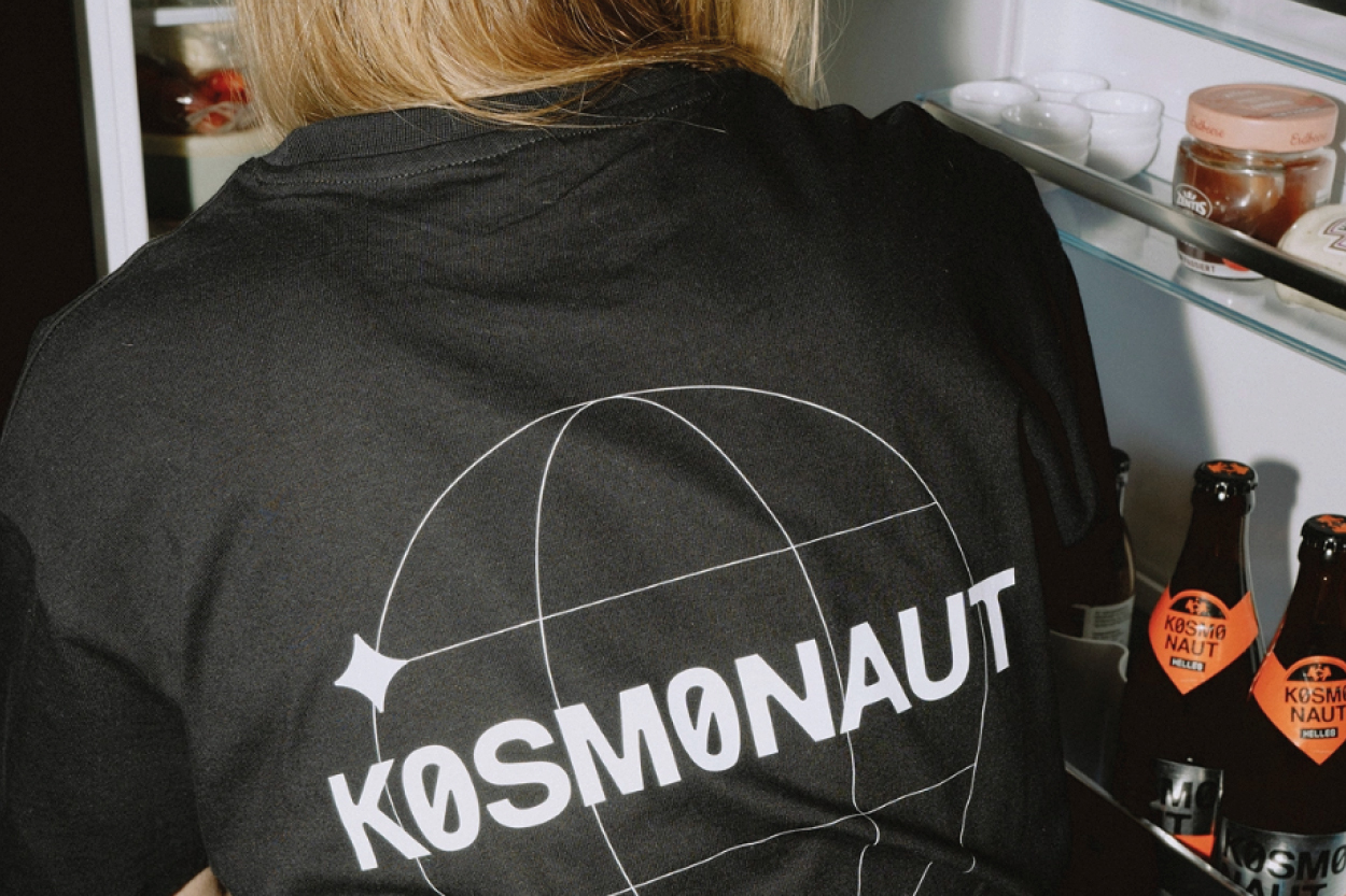 Eine Person steht mit dem Rücken zur Kamera vor einem geöffneten Kühlschrank. Auf ihrem schwarzen T-Shirt ist groß das Wort „KOSMONAUT“ sowie eine grafische Linienillustration zu sehen. Im Kühlschrank stehen Flaschen und Gläser, darunter mehrere Bierflaschen mit dem gleichen Branding.