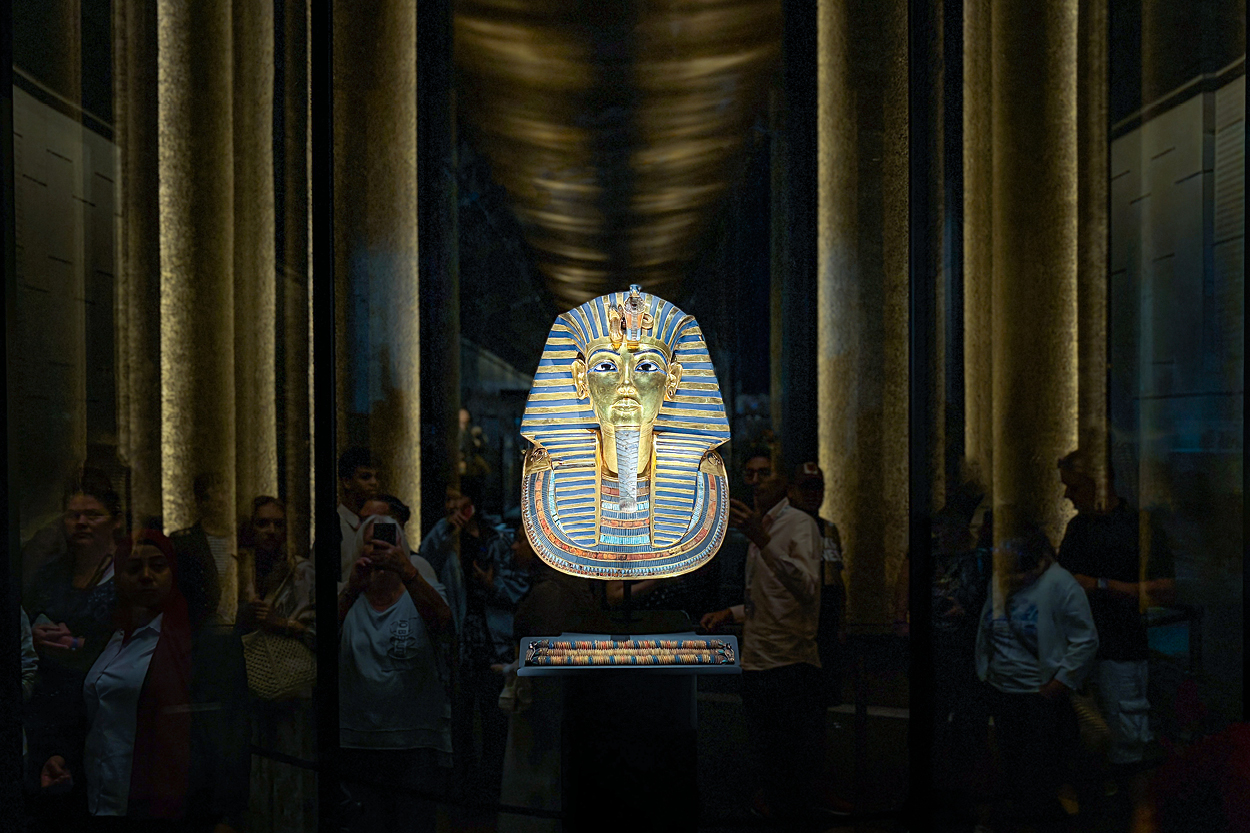 Eine goldene Totenmaske aus dem alten Ägypten wird in einer Glasvitrine in einem abgedunkelten Museumsraum ausgestellt. Die Maske ist zentral beleuchtet und zieht den Blick auf sich, während sich im Hintergrund Besucher und hohe Säulen spiegeln.