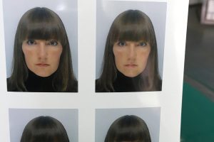Eine Hand mit rot lackierten Fingernägeln hält einen Bogen mit vier identischen Passfotos. Zu sehen ist eine Person mit schulterlan­gem, braunem Haar mit geradem Pony, blauen Augen und neutralem Gesichtsausdruck, gekleidet in einen schwarzen Rollkragenpullover. Hintergrund der Fotos ist hellgrau. Im Hintergrund des Gesamtbildes ist ein Arbeitsraum mit grüner Schneidematte erkennbar.