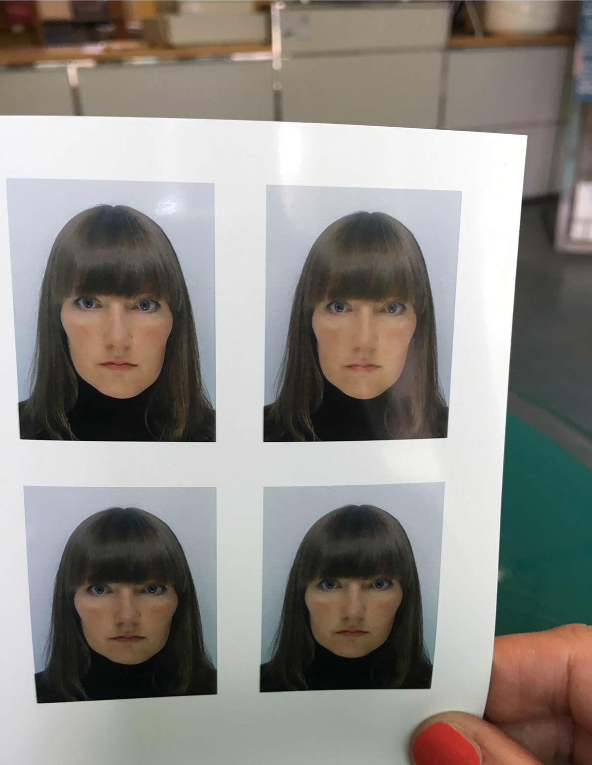 Eine Hand mit rot lackierten Fingernägeln hält einen Bogen mit vier identischen Passfotos. Zu sehen ist eine Person mit schulterlan­gem, braunem Haar mit geradem Pony, blauen Augen und neutralem Gesichtsausdruck, gekleidet in einen schwarzen Rollkragenpullover. Hintergrund der Fotos ist hellgrau. Im Hintergrund des Gesamtbildes ist ein Arbeitsraum mit grüner Schneidematte erkennbar.
