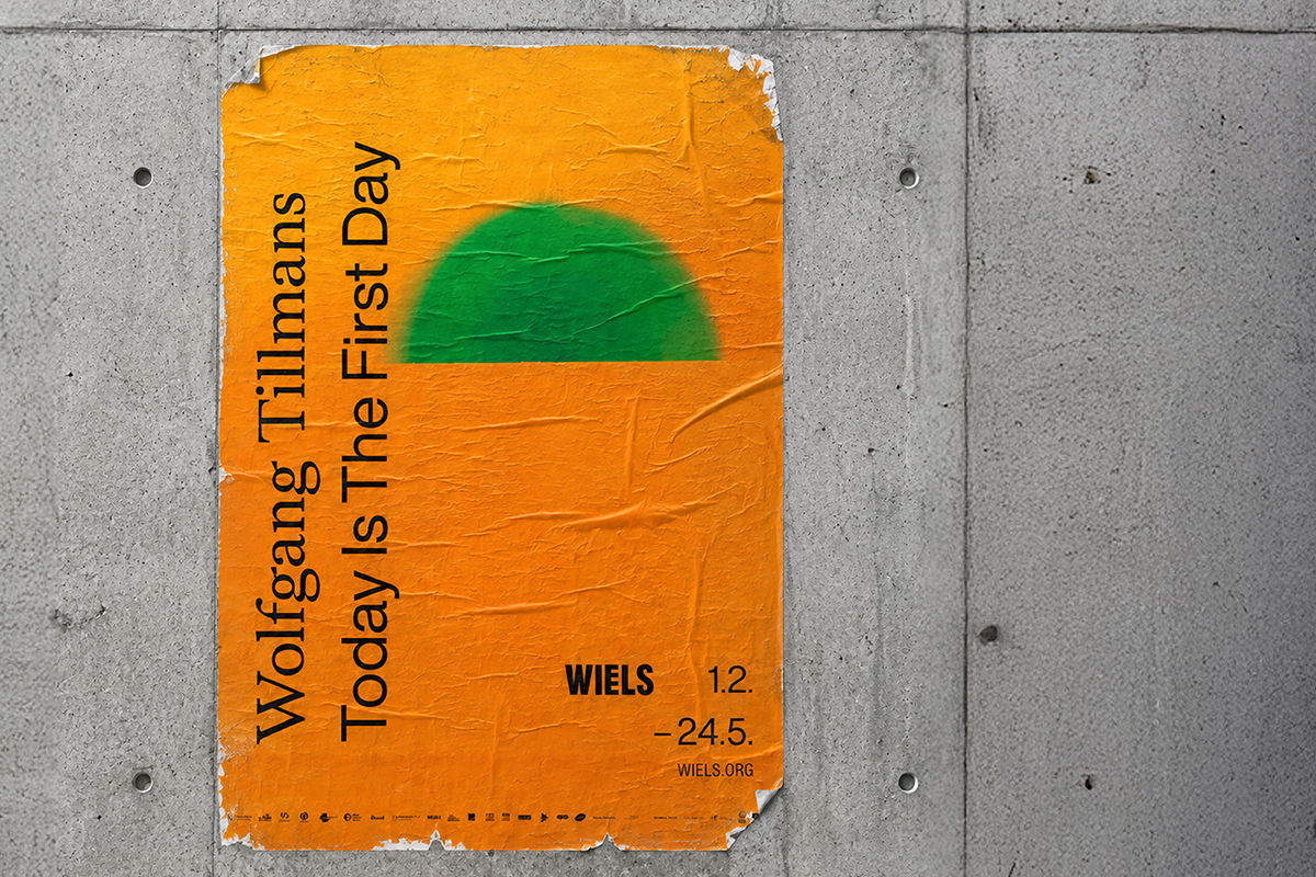 Nahaufnahme eines zerknitterten, an eine Betonwand geklebten Ausstellungsplakats. Leuchtend orangefarbenes Hochformat mit einem weichen grünen Halbkreis im oberen Drittel. Schwarzer Schriftzug in schräger Anordnung: „Wolfgang Tillmans – Today Is The First Day". Unten: „WIELS 1.2. – 24.5. wiels.org".