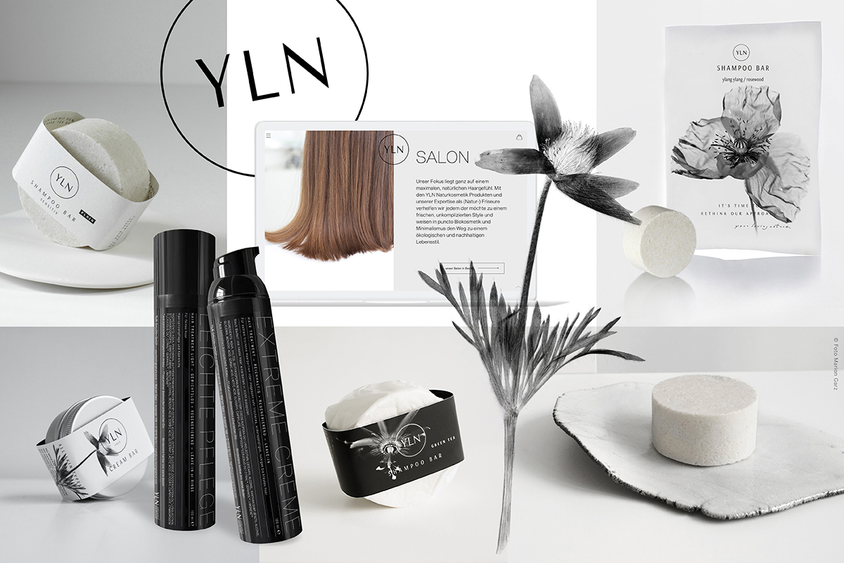 Produktcollage der Naturkosmetikmarke YLN auf weißem Hintergrund. Zu sehen sind: Shampoo Bars in Papierverpackung, zwei schwarze Sprühflaschen (Leichte Pflege, Extreme Crème), ein Tablet mit der YLN-Salon-Website, eine botanische Tuschezeichnung einer Blume, ein weißes Sachet „Shampoo Bar ylang ylang/rosewood" und ein runder Seifenblock auf Stein. Oben links das kreisförmige YLN-Logo.
