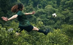 Baumwipfel, ganz in Grün, in denen eine junge Frau im Fußballoutfit einen Ball kickt