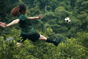 Baumwipfel, ganz in Grün, in denen eine junge Frau im Fußballoutfit einen Ball kickt
