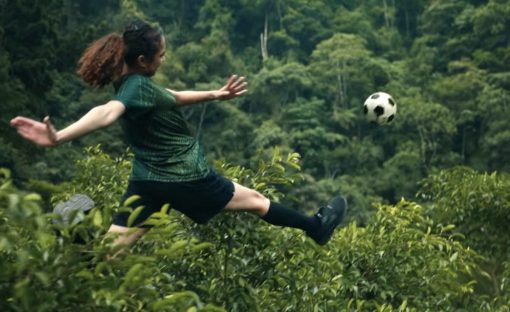 Baumwipfel, ganz in Grün, in denen eine junge Frau im Fußballoutfit einen Ball kickt