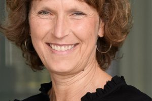 Porträt einer lächelnden Frau mit kurzen, leicht gewellten braunen Haaren. Sie trägt eine schwarze Bluse mit dezenten Rüschen am Kragen und an den Schultern sowie schlichte Creolen. Die Frau blickt direkt in die Kamera und wirkt freundlich, offen und selbstbewusst. Der Hintergrund ist unscharf gehalten und zeigt eine moderne Innenarchitektur mit Glas- und Betonelementen. Die Aufnahme vermittelt Professionalität und Nahbarkeit.