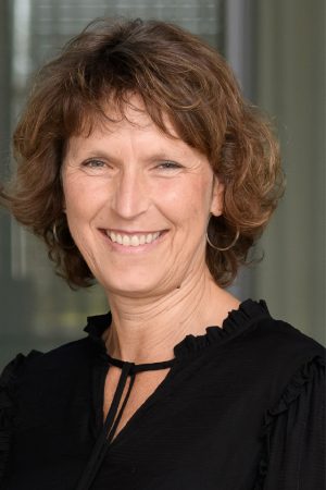 Porträt einer lächelnden Frau mit kurzen, leicht gewellten braunen Haaren. Sie trägt eine schwarze Bluse mit dezenten Rüschen am Kragen und an den Schultern sowie schlichte Creolen. Die Frau blickt direkt in die Kamera und wirkt freundlich, offen und selbstbewusst. Der Hintergrund ist unscharf gehalten und zeigt eine moderne Innenarchitektur mit Glas- und Betonelementen. Die Aufnahme vermittelt Professionalität und Nahbarkeit.