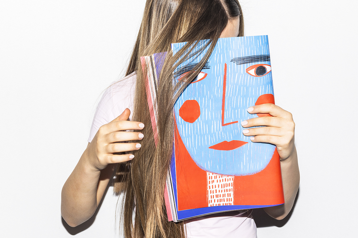 Mädchen, die Seite aus Vow Magazin mit illustriertem Gesicht in Hellblau und Rot vor ihr eigenes Gesicht hält. Drumherum ihre langen Haare