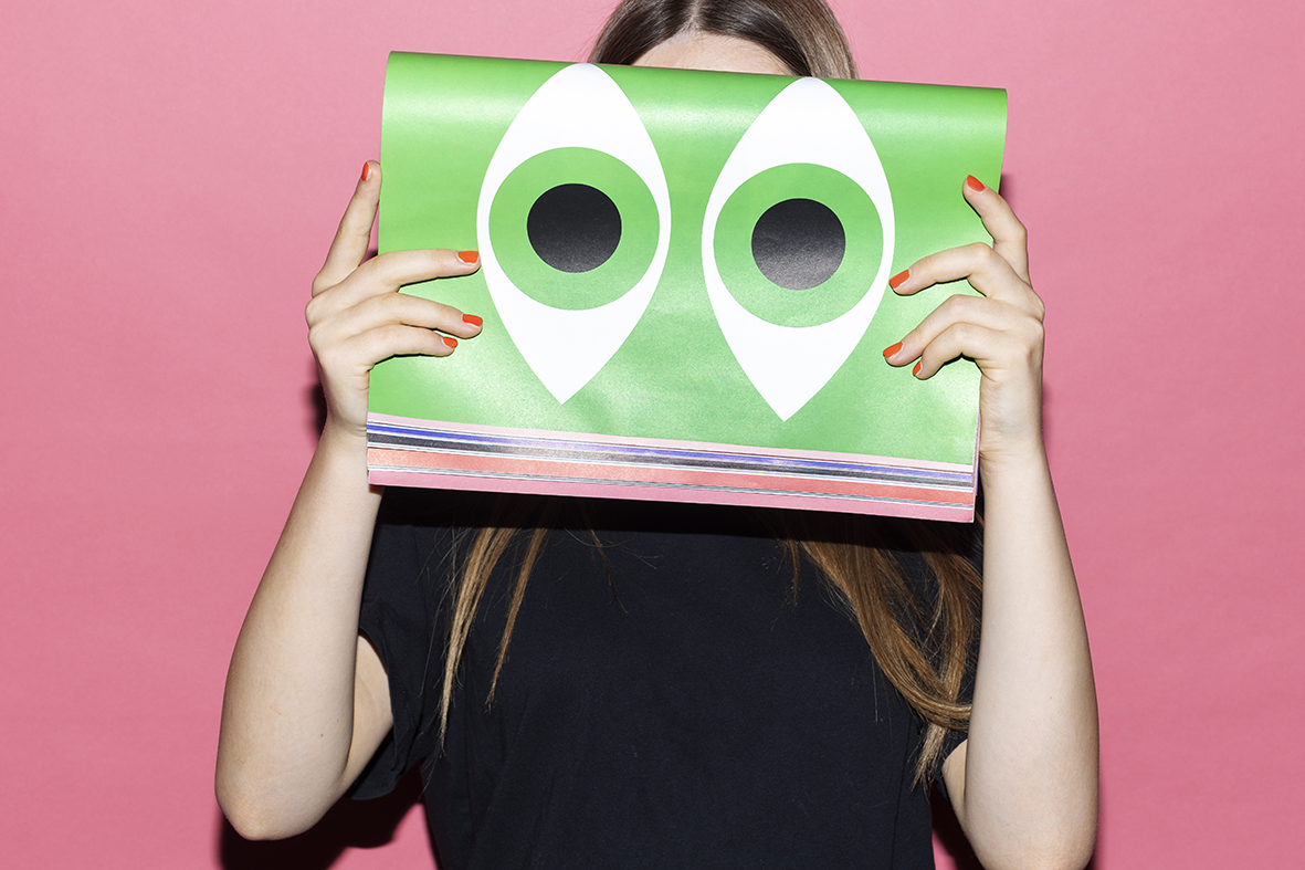 Mädchen, Kopf sieht man nicht, hält Magazin mit stilisiert illustrierten Augen in Grün vors Gesicht