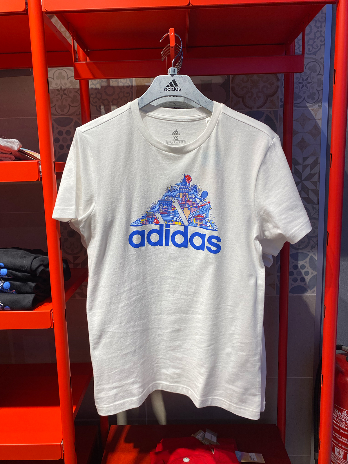 Weißes adidas-T-Shirt in Größe XS auf einem Bügel an einem roten Ladenregal, vor einer gemusterten Fliesenwand. Auf der Brust ein blaues adidas-Logo, dessen dreieckige Bergform mit einer detailreichen Illustration Istanbuler Motive gefüllt ist – Galata-Turm, Häuser und Wahrzeichen in Blau-, Rot- und Gelbtönen.