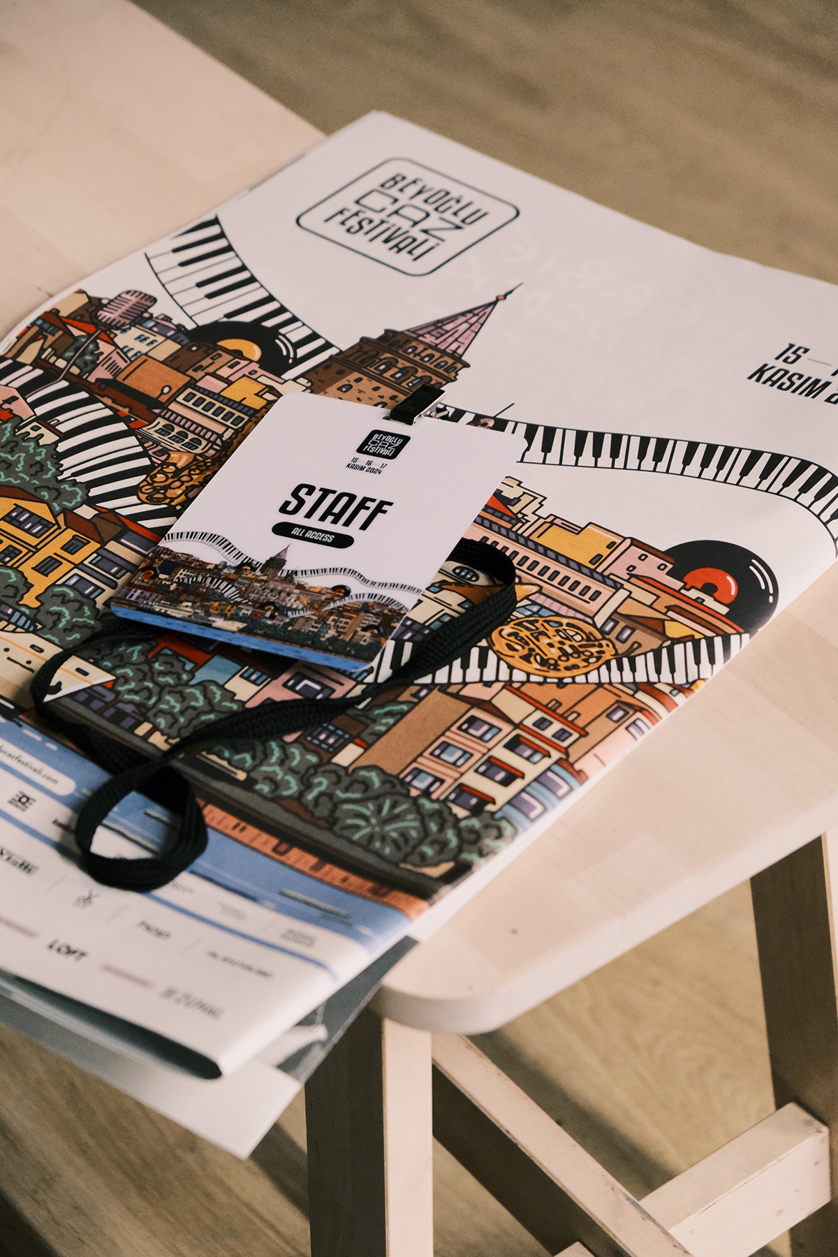 Fotografie eines Festival-Plakats auf einem hellen Holzhocker, darauf ein Staff-Akkreditierungsausweis mit schwarzem Lanyard. Das Plakat trägt den Schriftzug »BEYOĞLU CAZ FESTİVALİ« und zeigt eine dichte Illustration von Istanbuler Gebäuden und Wahrzeichen (u. a. Galata-Turm), verwoben mit Klaviertasten und einer Schallplatte. Der weiße Ausweis ist mit »STAFF – ALL ACCESS« und dem Datum »15 16 17 KASIM 2024« beschriftet.