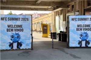 Großformatige Willkommensbanner markieren den Eingang zum NFC Summit 2026. Auf beiden Seiten des Eingangs stehen Plakate mit der Aufschrift „NFC Summit 2026 – Welcome / Bem Vindo“ sowie einer blauen Drachen-Maskottchenfigur im schwarzen Blazer. Dahinter öffnet sich ein überdachter Innenhof mit Industriearchitektur, gelber Infostele und Sitzgelegenheiten im Freien. Die Aufnahme vermittelt den Eingangsbereich und die Atmosphäre des Veranstaltungsortes.