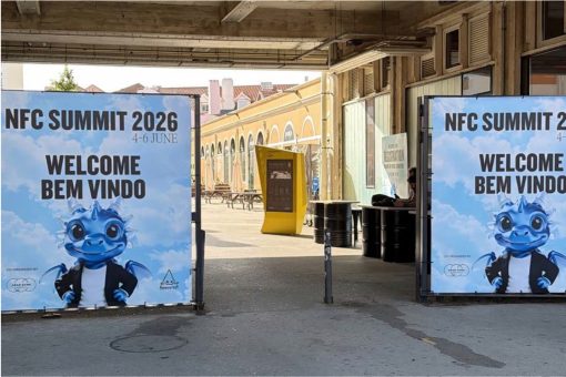 Großformatige Willkommensbanner markieren den Eingang zum NFC Summit 2026. Auf beiden Seiten des Eingangs stehen Plakate mit der Aufschrift „NFC Summit 2026 – Welcome / Bem Vindo“ sowie einer blauen Drachen-Maskottchenfigur im schwarzen Blazer. Dahinter öffnet sich ein überdachter Innenhof mit Industriearchitektur, gelber Infostele und Sitzgelegenheiten im Freien. Die Aufnahme vermittelt den Eingangsbereich und die Atmosphäre des Veranstaltungsortes.