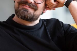 Simon Graff sitzt entspannt auf einem senfgelben Sofa und lächelt direkt in die Kamera. Er trägt ein schwarzes Langarmshirt, eine runde schwarze Brille sowie eine Smartwatch und stützt den Kopf lässig auf die rechte Hand. Der helle, unscharfe Hintergrund lenkt den Fokus auf das freundliche Porträt.