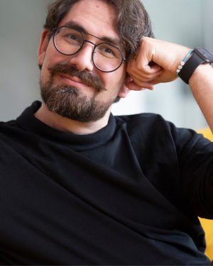 Simon Graff sitzt entspannt auf einem senfgelben Sofa und lächelt direkt in die Kamera. Er trägt ein schwarzes Langarmshirt, eine runde schwarze Brille sowie eine Smartwatch und stützt den Kopf lässig auf die rechte Hand. Der helle, unscharfe Hintergrund lenkt den Fokus auf das freundliche Porträt.