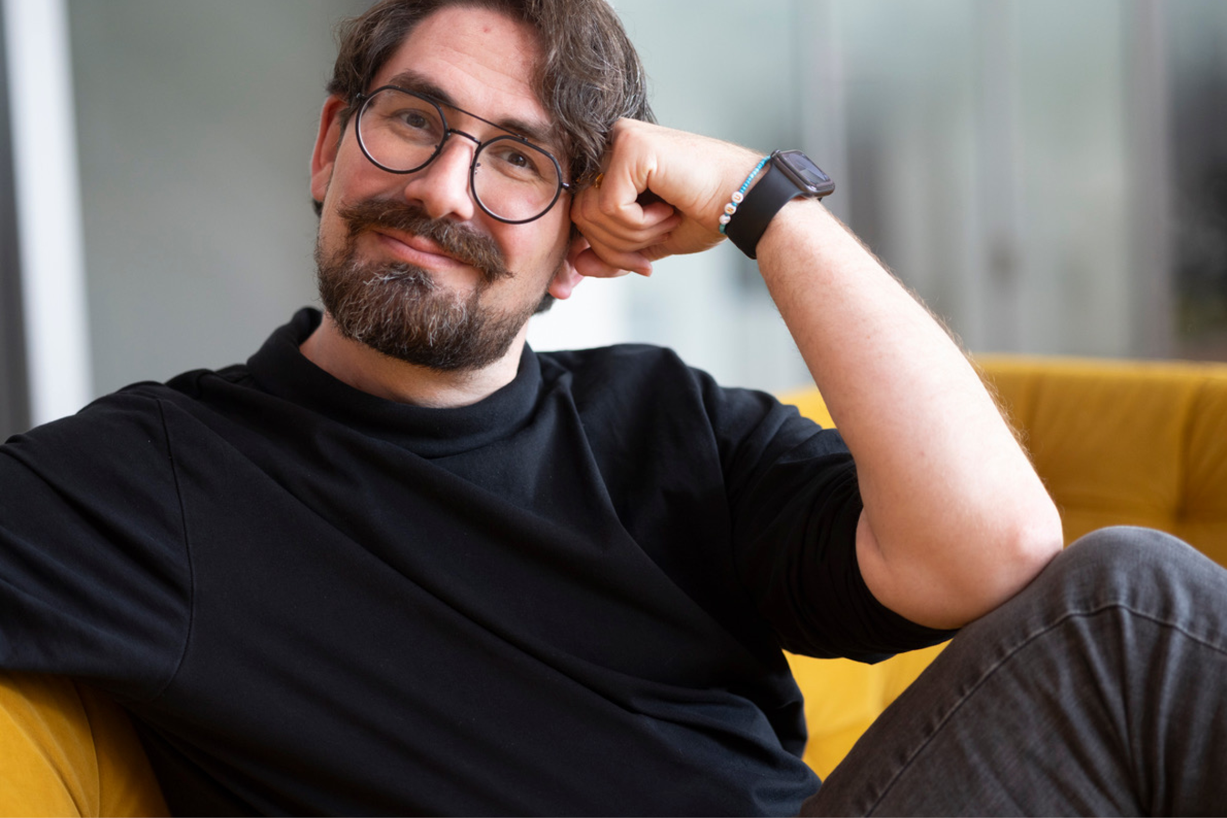 Simon Graff sitzt entspannt auf einem senfgelben Sofa und lächelt direkt in die Kamera. Er trägt ein schwarzes Langarmshirt, eine runde schwarze Brille sowie eine Smartwatch und stützt den Kopf lässig auf die rechte Hand. Der helle, unscharfe Hintergrund lenkt den Fokus auf das freundliche Porträt.