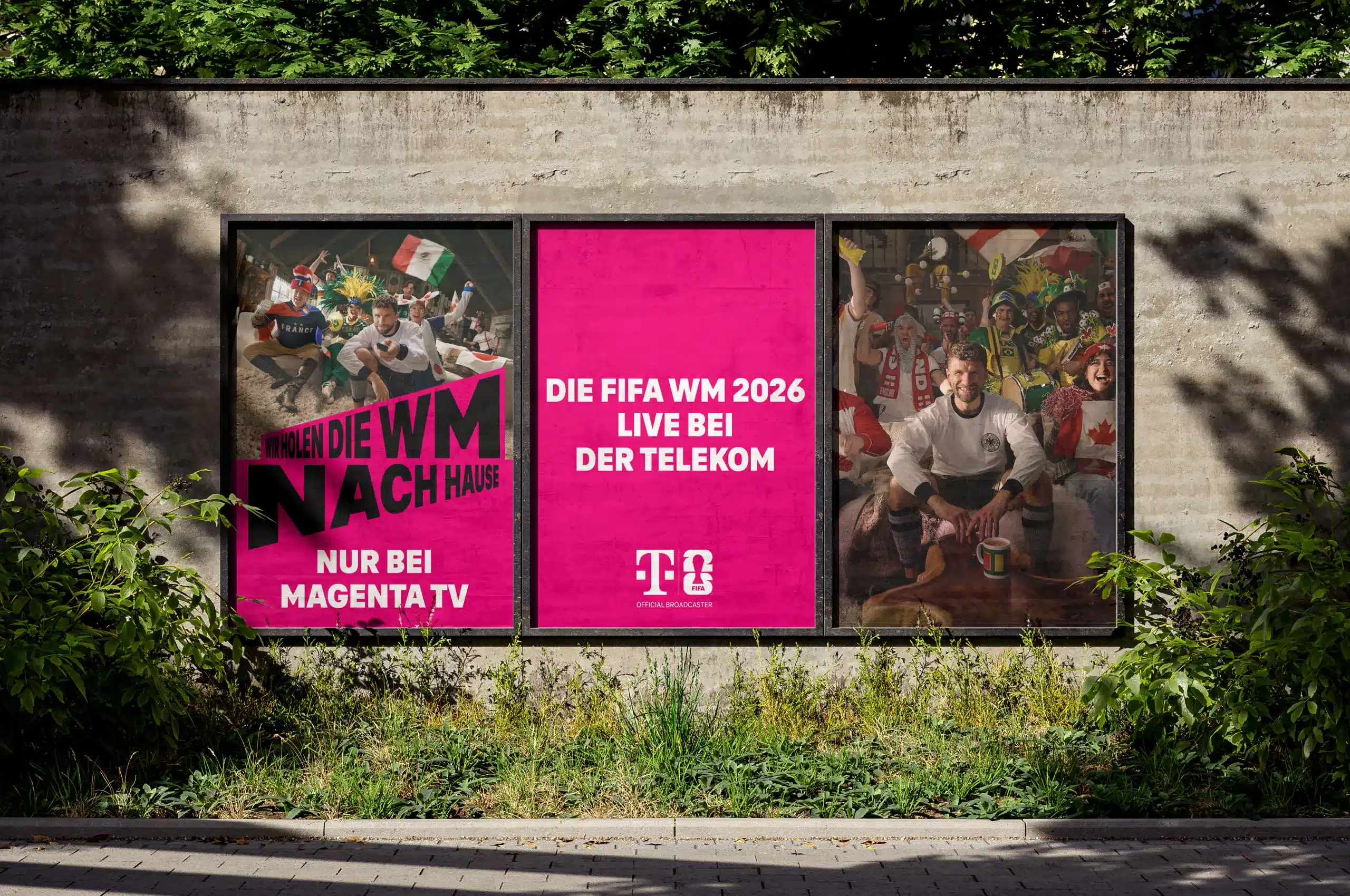 Drei großformatige Plakatflächen an einer Betonwand bewerben die FIFA-Weltmeisterschaft 2026 auf MagentaTV. Das mittlere, vollständig magentafarbene Plakat trägt den weißen Schriftzug „Die FIFA WM 2026 live bei der Telekom“ sowie das Telekom-Logo. Links und rechts zeigen Motive mit jubelnden Fußballfans aus verschiedenen Nationen; im Vordergrund sitzt jeweils ein Fan im Trikot. Auf dem linken Plakat steht zusätzlich in großer Typografie „Wir holen die WM nach Hause – nur bei MagentaTV“. Die Plakate sind von Grünpflanzen und Bäumen eingerahmt und wirken wie eine aufmerksamkeitsstarke Out-of-Home-Kampagne.