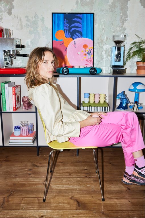 Eine Person sitzt seitlich auf einem gelben Stuhl in einem kreativ eingerichteten Studio oder Wohnraum und blickt direkt in die Kamera. Sie trägt eine cremefarbene Bluse, eine leuchtend pinke Hose, rosa Socken und mehrfarbige Sneaker. Hinter ihr steht ein offenes Regal mit Designobjekten, Büchern, Keramik und einer Espressomaschine sowie einer Kaffeemühle. An der Wand hängt ein farbenfrohes Gemälde in kräftigen Blau-, Rosa- und Orangetönen. Die Kombination aus Designobjekten, Kunst und kräftigen Farben vermittelt eine moderne, kreative Atelieratmosphäre.