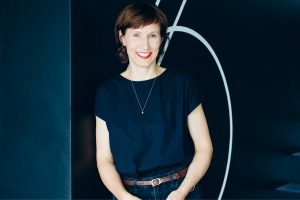 Porträt einer lächelnden Frau mit kurzen braunen Haaren vor einem dunklen Hintergrund mit grafischen weißen Linien. Sie trägt ein schwarzes Kurzarmshirt, dunkle Jeans mit braunem Gürtel sowie dezenten Goldschmuck und blickt direkt in die Kamera.