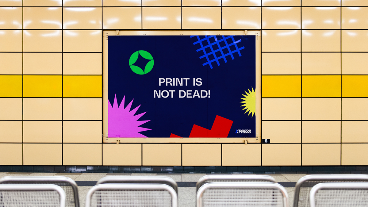 U-Bahn-Plakat auf hellgelbem Fliesenhintergrund. Dunkelblau mit Text »PRINT IS NOT DEAD!« in Weiß, XPRESS-Logo, grüner Kreis, magenta und gelbe Strahlen, rote Pfeile.