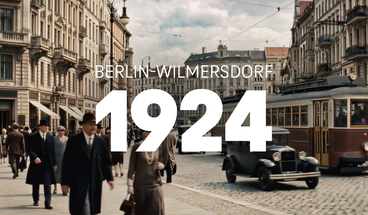 Historische Straßenszene im Berlin-Wilmersdorf des Jahres 1924 mit elegant gekleideten Passant:innen, einem Oldtimer und einer Straßenbahn vor prächtigen Altbaufassaden. Im Vordergrund steht in großer weißer Schrift „1924“, darüber „Berlin-Wilmersdorf“.