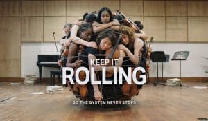 Eine Gruppe junger Musiker:innen mit Geigen und Bratschen ist in einem Proberaum eng ineinander verschlungen und bildet eine kugelförmige Komposition. Im Vordergrund steht der Schriftzug „KEEP IT ROLLING“, darunter „SO THE SYSTEM NEVER STOPS“.